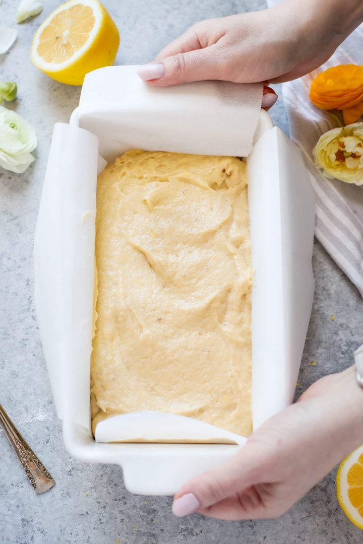 Hands holding a parchment-lined loaf pan filled with smooth banana lemon bread batter, ready for the oven. A lemon and fresh flowers frame the shot, adding a touch of brightness to this banana bread recipe with lemon juice.