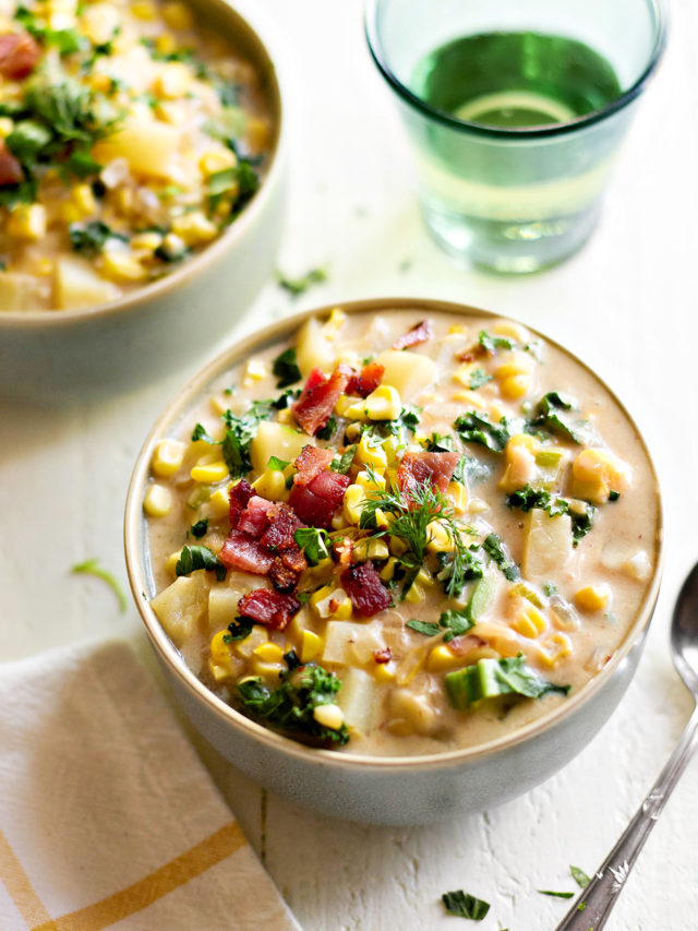 A close-up of bacon potato corn chowder served in a bowl on a white wooden table. The creamy broth is filled with tender potatoes, sweet roasted corn, and crispy bacon, garnished with fresh parsley and dill. A spoon rests beside the bowl, ready for serving.
