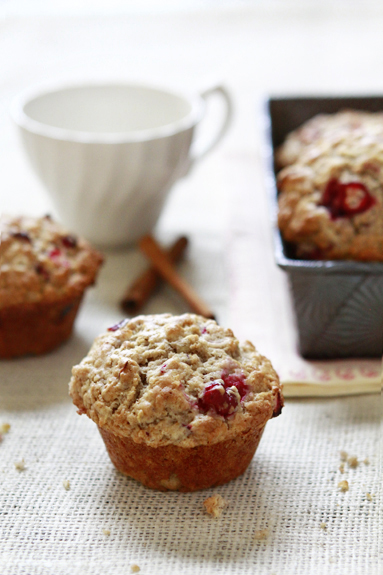 Two apple cranberry oatmeal muffins, with a tea cup and a loaf tin in the background. 