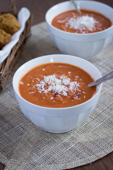 Two bowls of vegan tomato bisque, with a basket of rolls in the background.