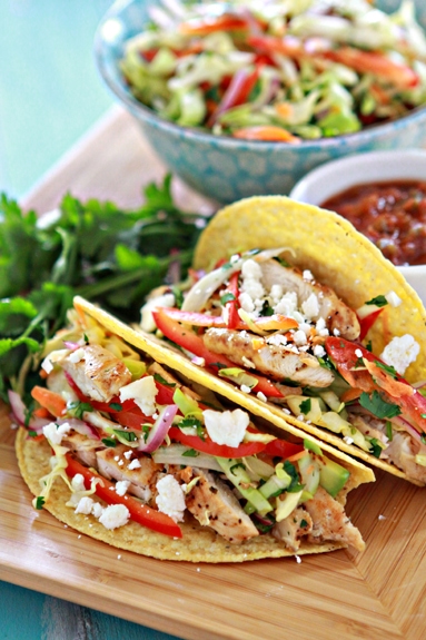 a photo of Easy Lemon Pepper Chicken Tacos with Slaw on a bamboo tray with bowls of cilantro lime slaw and salsa in the background