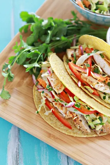 a photo of Easy Lemon Pepper Chicken Tacos with Slaw on a bamboo tray with bowls of cilantro lime slaw in the background