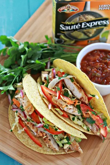 a photo of Easy Lemon Pepper Chicken Tacos with Slaw on a bamboo tray with bowl of salsa in the background