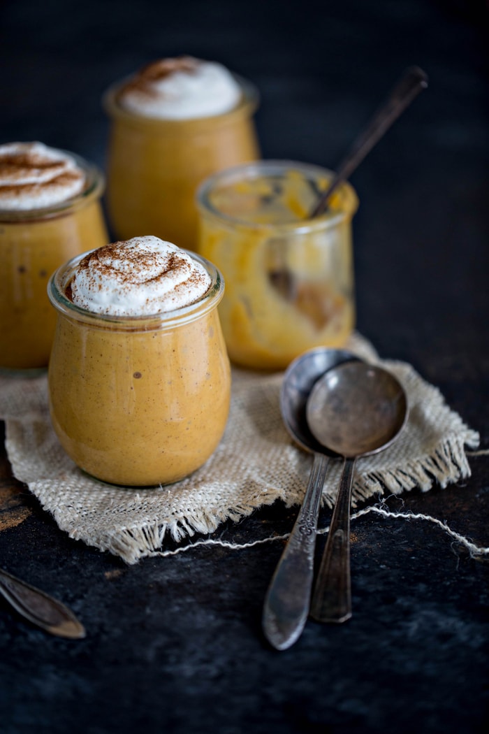 Four jars, three of which are filled with pumpkin spice pudding and garnished with whipped cream. The fourth jar has been eaten. Two spoons rest in the foreground. 