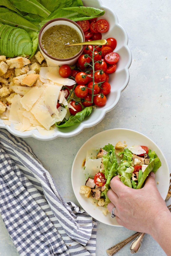 A hand is filling romaine leaves with Caesar salad ingredients—grilled chicken, cherry tomatoes, parmesan, and croutons—on a white plate. A larger serving dish nearby shows the full deconstructed salad with dressing.