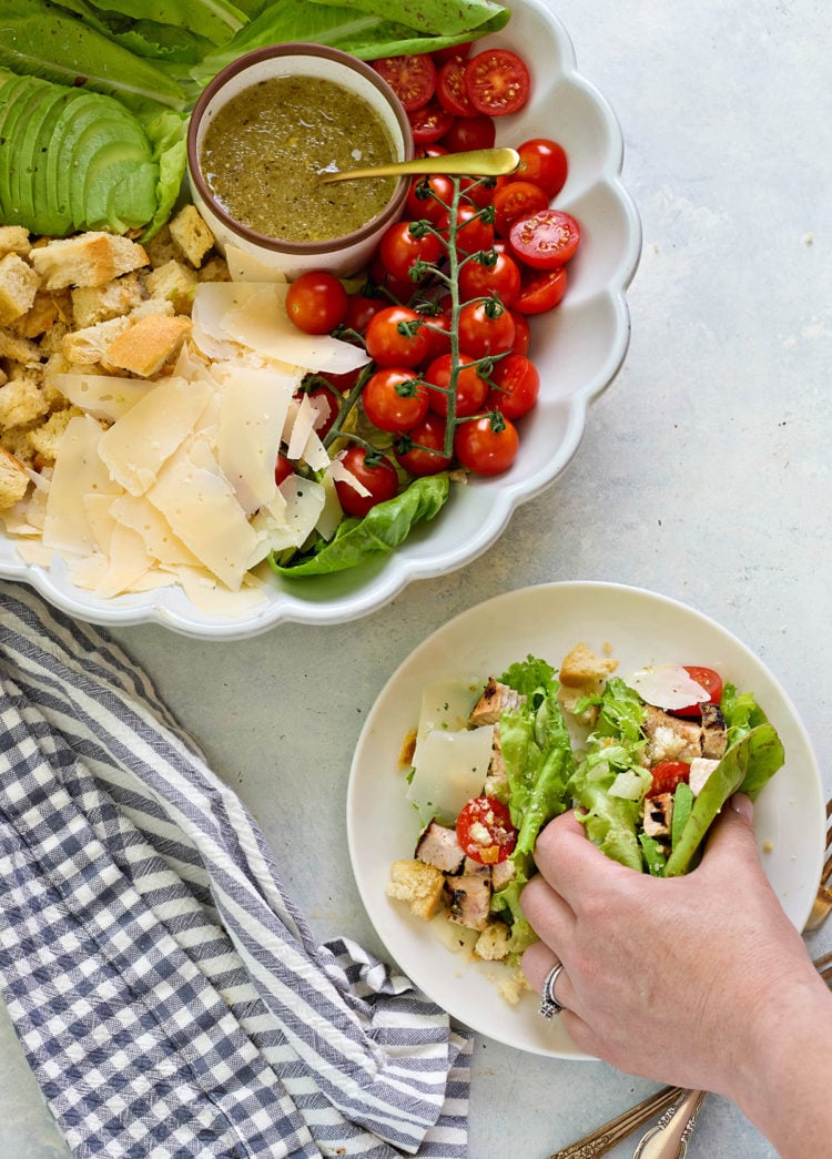 A hand is filling romaine leaves with Caesar salad ingredients—grilled chicken, cherry tomatoes, parmesan, and croutons—on a white plate. A larger serving dish nearby shows the full deconstructed salad with dressing.