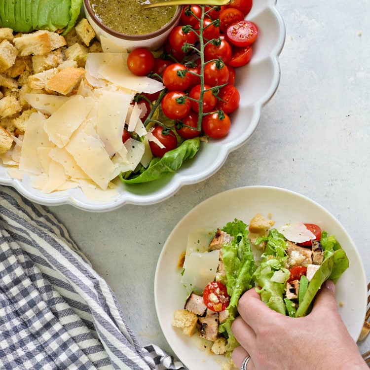 A hand is filling romaine leaves with Caesar salad ingredients—grilled chicken, cherry tomatoes, parmesan, and croutons—on a white plate. A larger serving dish nearby shows the full deconstructed salad with dressing.