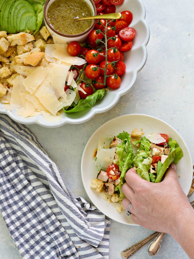 A hand is filling romaine leaves with Caesar salad ingredients—grilled chicken, cherry tomatoes, parmesan, and croutons—on a white plate. A larger serving dish nearby shows the full deconstructed salad with dressing.