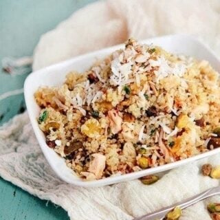 a white bowl filled with pineapple quinoa salad that is garnished with toasted coconut. the bowl sits on top of a turquoise wooden background with a white kitchen towel, fork, and pistachios sprinkled around it