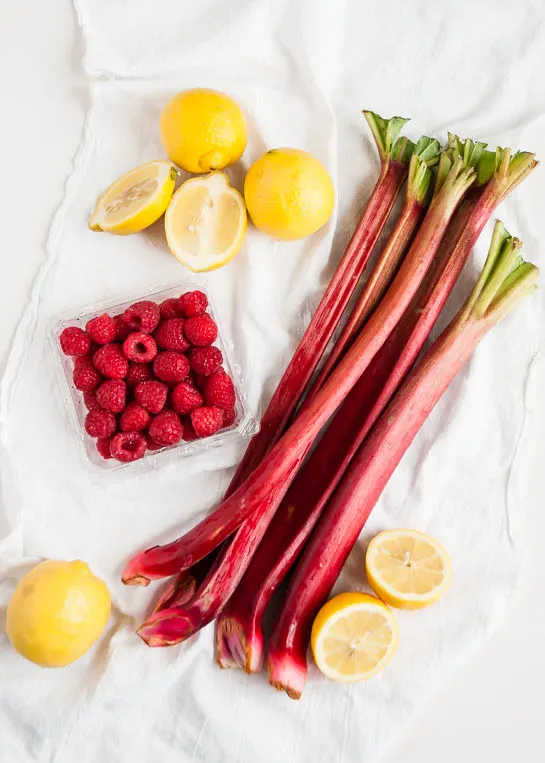 ingredients to make rhubarb lemonade - raspberries, lemons, and rhubarb on a white tablecloth