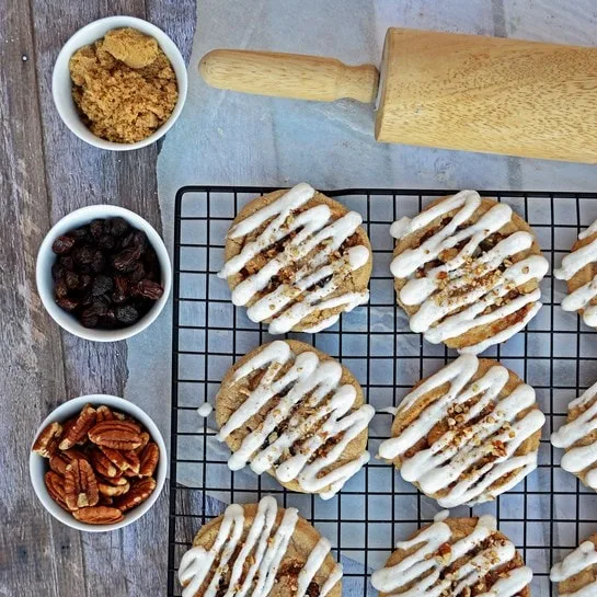 Overhead view of cinnamon roll cookies on a wire rack. Small bowls of pecans, raisins, and brown sugar lay nearby, as does a wood rolling pin. 