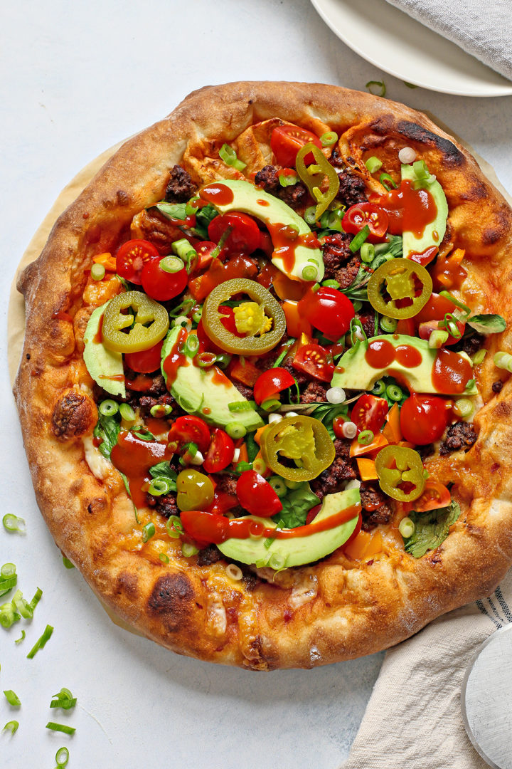 a taco pizza with hot sauce, avocado, and pickled jalapenos on top