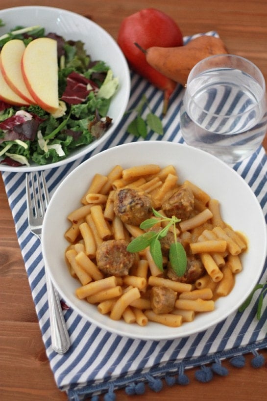 a bowl of meatballs and pasta with butternut squash sauce on a wooden table with a bowl of salad