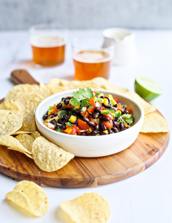A white ceramic bowl filled with black bean salad sits on a round wooden board surrounded by tortilla chips. The salad includes black beans, corn, tomatoes, peppers, and cilantro. A halved lime and two glasses of amber-colored beer are visible in the background.