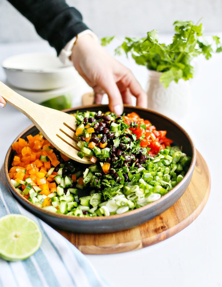 A large ceramic bowl filled with fresh ingredients for black bean salad. A wooden spatula is mid-motion, mixing layers of chopped orange bell pepper, cucumber, tomato, cilantro, green onion, and black beans. A halved lime and a bunch of fresh cilantro sit nearby.