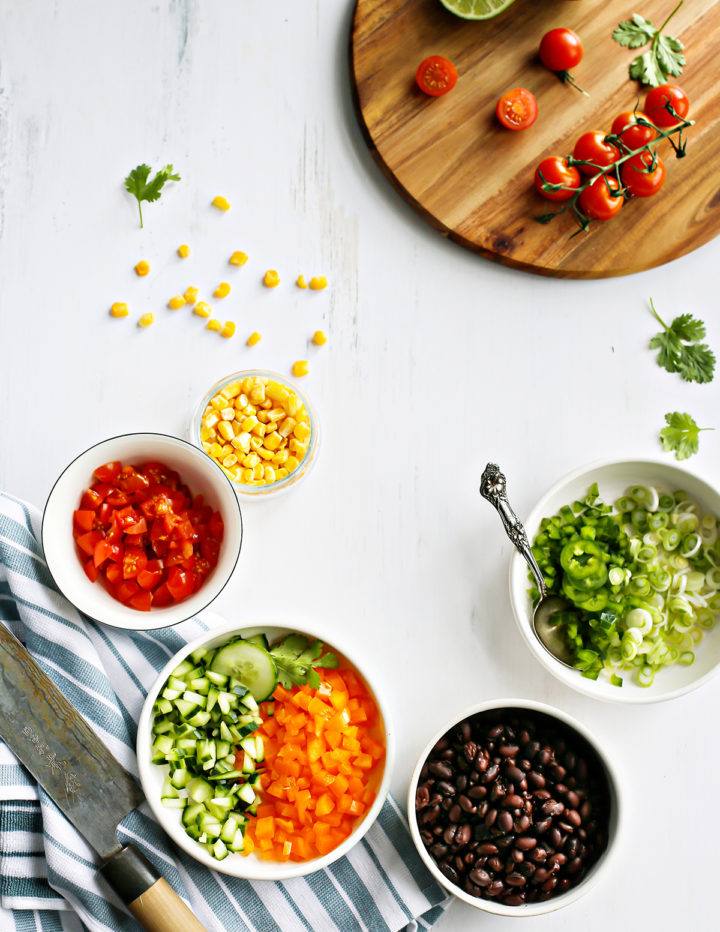 An overhead view of prepped black bean salad ingredients in small bowls on a white table. Bowls include diced tomatoes, corn, cucumber, orange bell pepper, black beans, and green onion. A striped kitchen towel, chef’s knife, and cutting board with tomatoes are also visible.