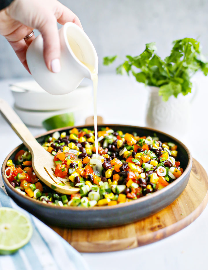 A hand pours lime dressing from a small white pitcher over a large bowl of black bean and corn salad. A wooden spoon rests in the salad, and the ingredients are bright and colorful with lots of chopped vegetables and beans.