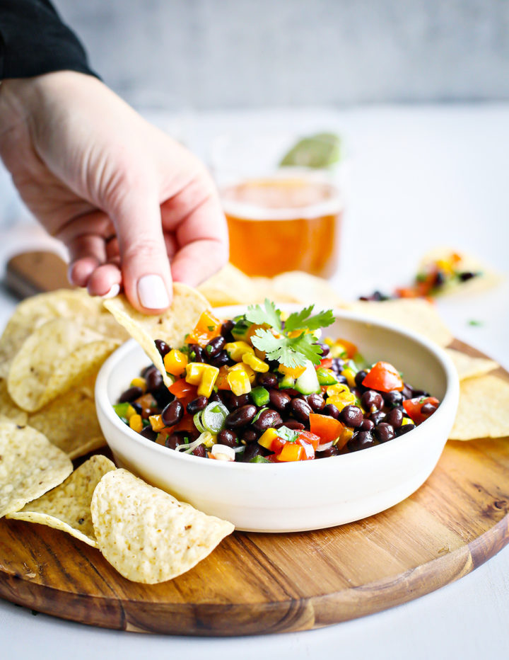 A hand with light nail polish scoops black bean salad with a tortilla chip from a white bowl. The bowl rests on a wooden board with more chips around it. The colorful salad features black beans, corn, chopped peppers, tomatoes, green onions, and a cilantro garnish.