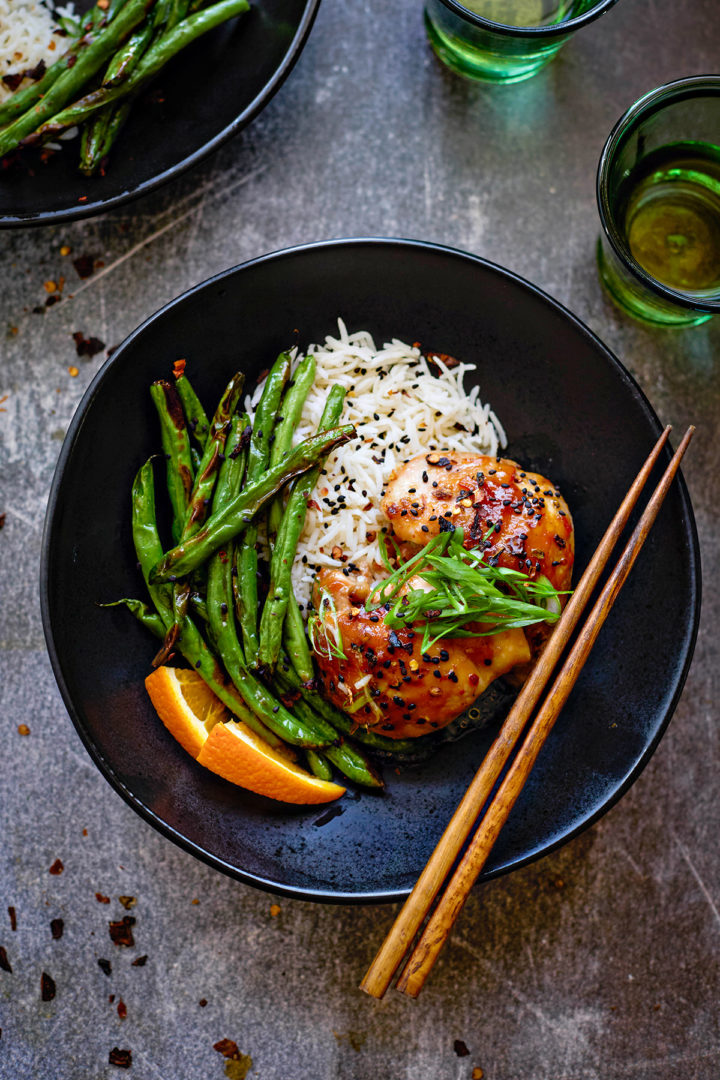 A bowl of steamed white rice topped with orange chicken and orange marmalade sauce, with chopsticks placed nearby. The sauce glistens on the chicken and adds a deep orange hue. 