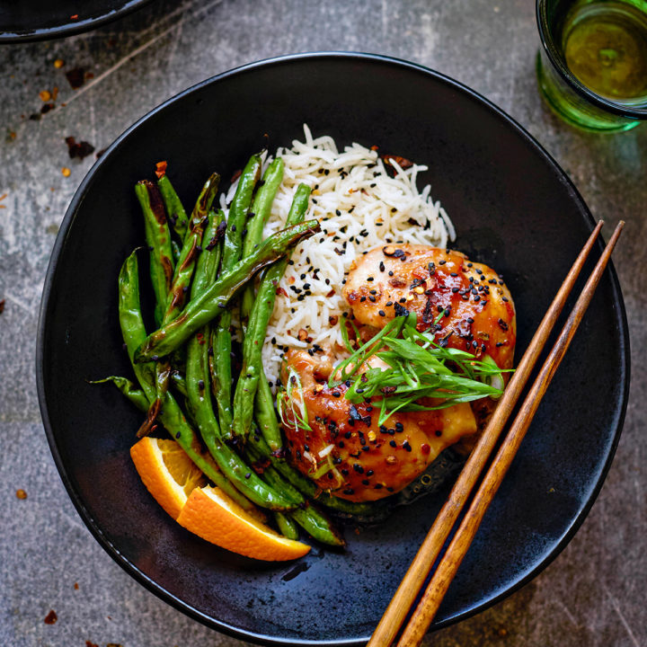 Flat lay view of orange marmalade chicken in a bowl with rice, green beans, and sliced fresh oranges. The chicken is garnished with thinly sliced green onions and sesame seeds. The marmalade sauce clings to each piece of chicken with a sticky, slightly thickened texture.