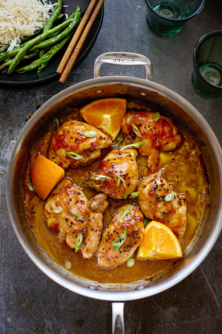 A clean, styled photo of cooked orange marmalade chicken in a skillet, with fresh orange slices and chopped green onions around the edge. The orange marmalade chicken sauce has thickened into a rich glaze that clings to each chicken piece, perfect for a quick weeknight meal.