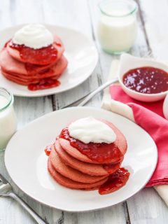 two white plates with stacks of pink pancakes on them served with strawberry jam and whipped cream