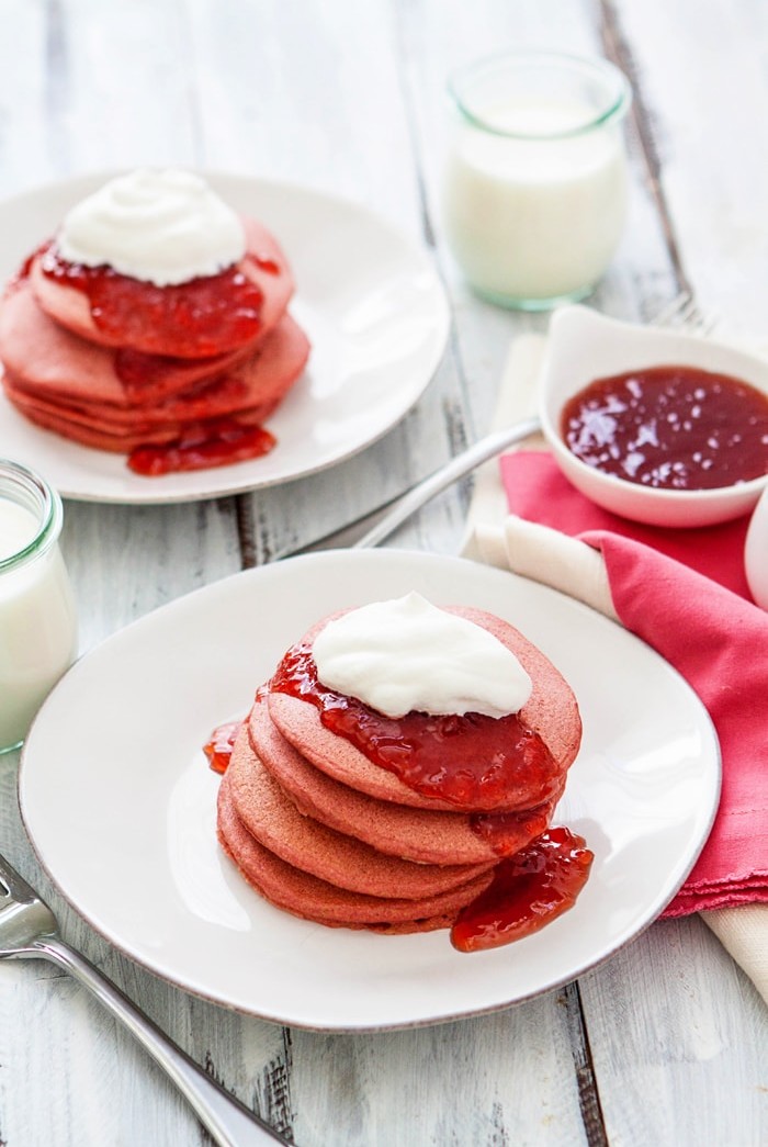 two white plates with stacks of pink pancakes on them served with strawberry jam and whipped cream
