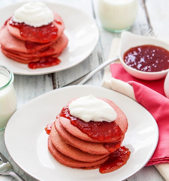 two white plates with stacks of pink pancakes on them served with strawberry jam and whipped cream