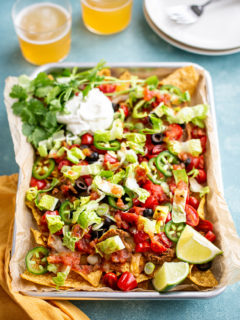 photo of nachos recipe on a sheet pan with sour cream and cilantro garnishes, next to two glasses of beer
