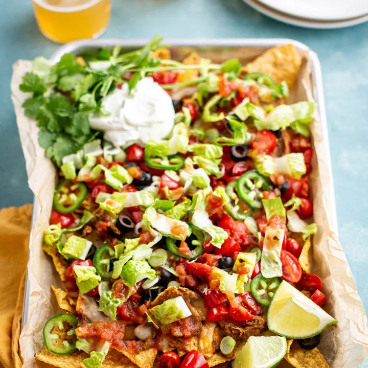 photo of nachos recipe on a sheet pan with sour cream and cilantro garnishes, next to two glasses of beer