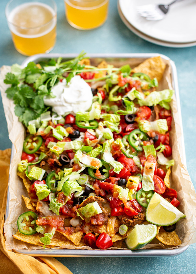 photo of nachos recipe on a sheet pan with sour cream and cilantro garnishes, next to two glasses of beer