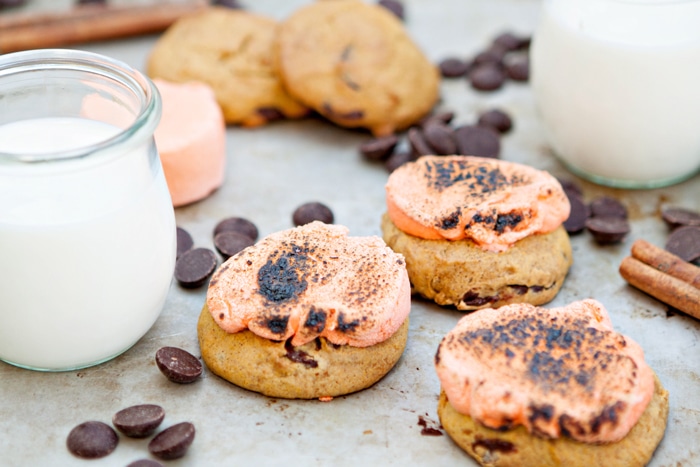 Three pumpkin spice chocolate chip cookies topped with toasted marshmallows, next to a glass of milk. Chocolate chips are scattered around the cookies. 