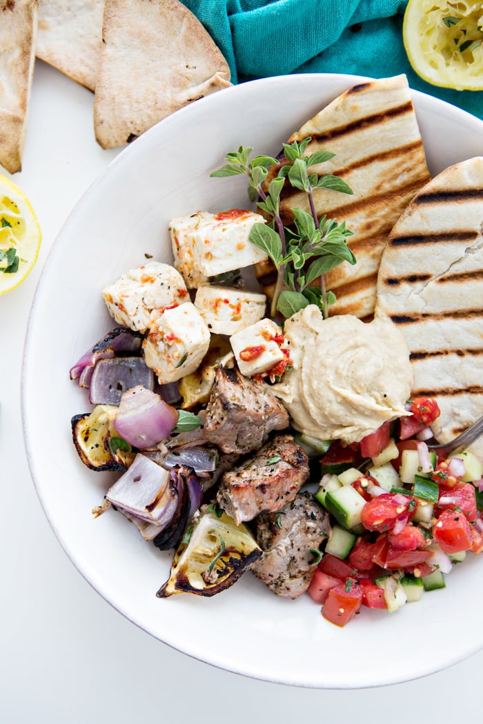 a delicious greek recipe of grilled greek pork tenderloin in a white bowl with tomato and cucumber salad, feta, hummus, and pita bread