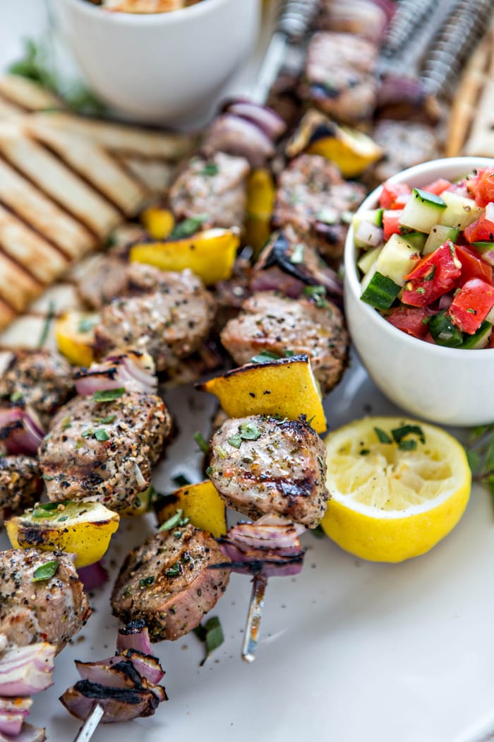 photo of greek pork kebabs on skewers with lemon and red onion being served on a white platter