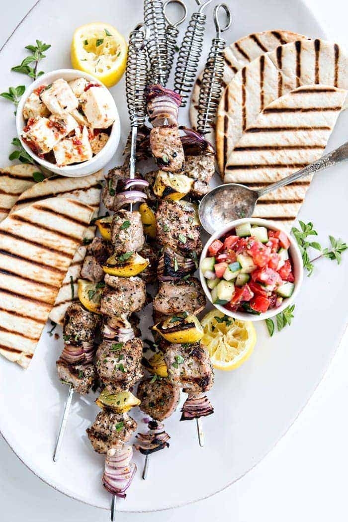 overhead photo of a kebab platter with pork on skewers, a bowl of tomato cucumber salad, grilled pita bread, and marinated feta