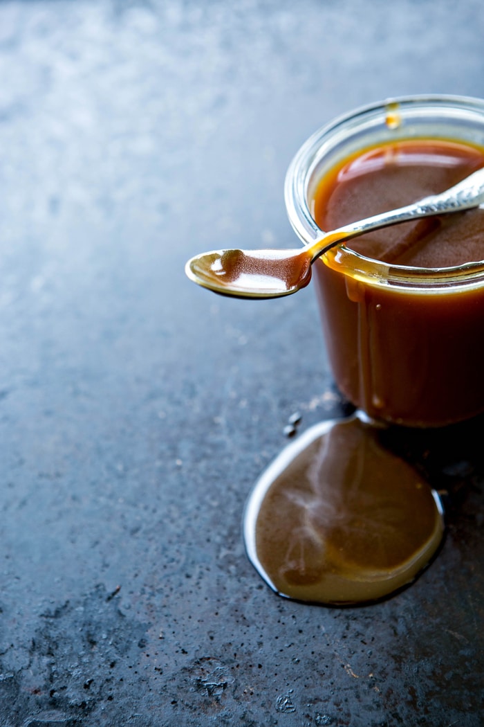 photo of a jar of homemade bourbon caramel sauce with a spoon sitting on top and caramel dripping down the side