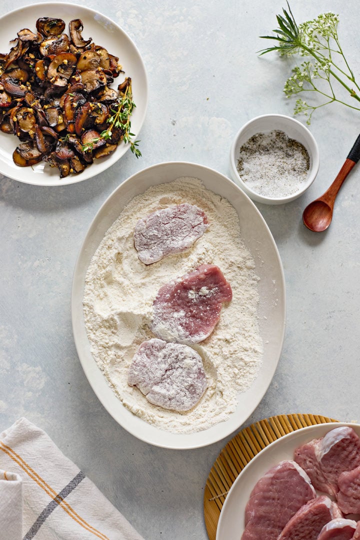Overhead view of a light blue countertop with four food prep elements arranged neatly. In the center, an oval dish holds flour with three raw pork medallions partially coated in it. To the top left, a white plate is filled with sautéed sliced mushrooms and a few sprigs of fresh flowers. To the top right, a small white bowl contains a coarse salt and pepper mix, with a wooden spoon beside it. In the bottom right corner, a plate of raw, uncoated pork medallions is partially visible, and a folded dish towel with yellow and gray stripes sits in the lower left corner.