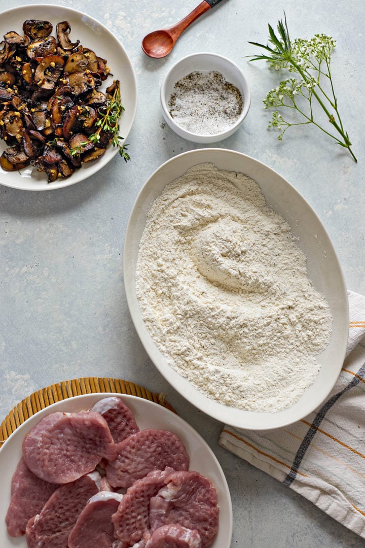 Overhead view of ingredients for making pork marsala with mushrooms. In the center is an oval dish filled with flour for dredging. At the bottom left, a plate of raw pork tenderloin medallions sits partially in frame. At the top left, a bowl of sautéed mushrooms is garnished with sprigs of thyme. A ramekin of salt and pepper is placed above the flour, next to a wooden spoon. A striped kitchen towel and a sprig of flowering herb are also visible on the light blue background.
