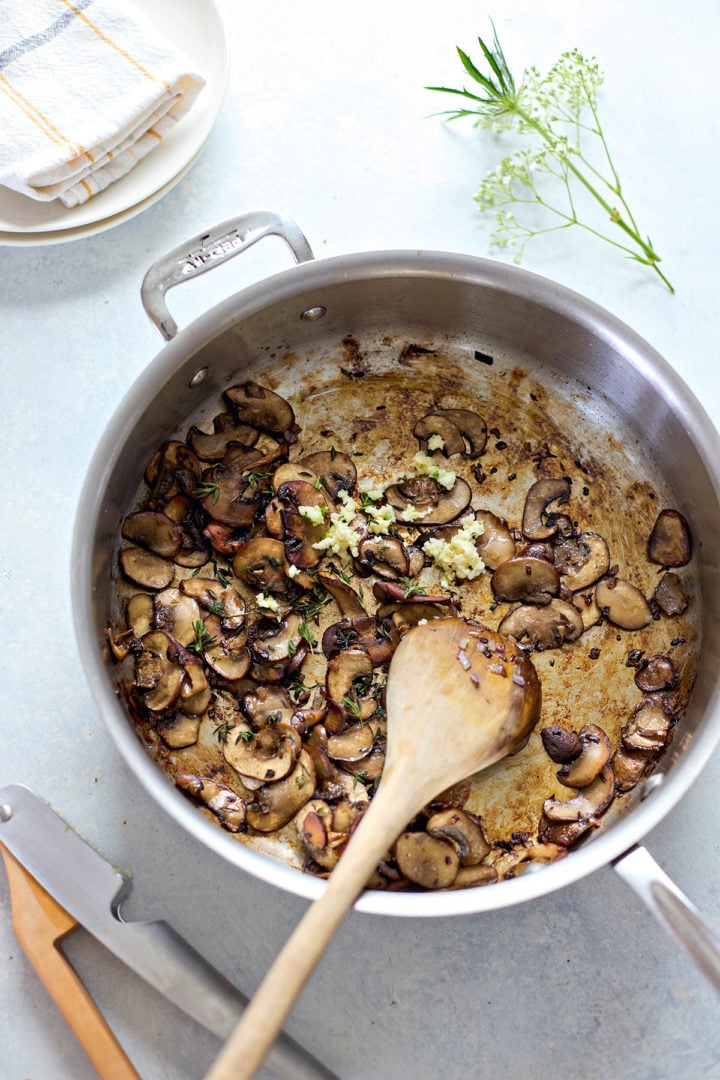 Overhead view of a stainless steel pan with sautéed mushrooms and minced garlic being cooked for pork marsala with mushrooms. The sliced mushrooms are browned and pushed to one side of the pan, with a pile of freshly added garlic in the center. Small pieces of chopped shallot and sprigs of thyme are scattered throughout. A wooden spoon rests in the pan, and part of a knife and folded dish towel are visible in the background.

