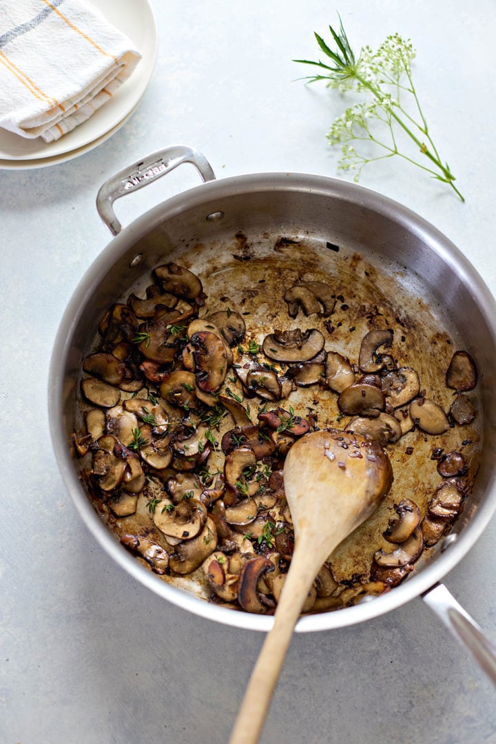 Sautéed mushrooms in a stainless steel skillet, shown from above as part of preparing marsala pork tenderloin. The mushrooms are browned and glossy, with bits of caramelized shallot and sprigs of fresh thyme scattered throughout. A wooden spoon rests in the pan, and browned fond is visible on the bottom. A folded cloth napkin and flowering herb sprig are partially visible in the background.