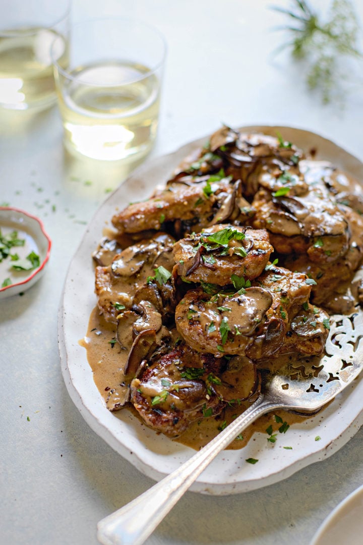 A white oval platter filled with pork tenderloin marsala. The seared pork medallions are coated in a creamy brown mushroom sauce and garnished with freshly chopped parsley. Sliced mushrooms are visible throughout the sauce. A serving spoon rests on the platter, and two glasses of white wine sit in the background along with a small dish of extra parsley.