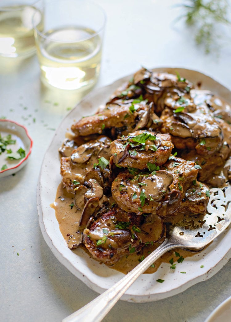 Overhead view of a serving platter filled with seared pork medallions topped with marsala sauce for pork. The creamy mushroom sauce is generously spooned over the meat and garnished with chopped parsley. A silver serving spoon rests on the platter. Two glasses of white wine and a small dish of additional parsley sit nearby on the light-colored surface.