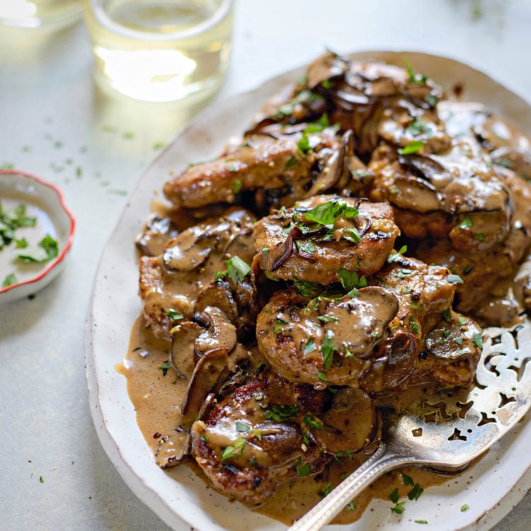 Overhead view of a serving platter filled with seared pork medallions topped with marsala sauce for pork. The creamy mushroom sauce is generously spooned over the meat and garnished with chopped parsley. A silver serving spoon rests on the platter. Two glasses of white wine and a small dish of additional parsley sit nearby on the light-colored surface.