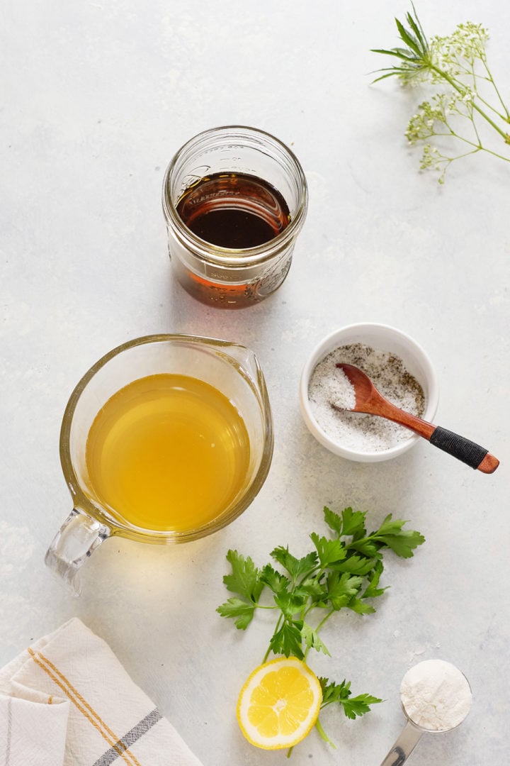 Overhead view of ingredients used to make marsala sauce for pork. A small glass measuring cup of chicken broth and a jar of Marsala wine are placed side by side on a light surface. Nearby are a ramekin of salt and pepper with a wooden spoon, a lemon half, fresh parsley leaves, and a tablespoon of flour. A folded dish towel with stripes is partially visible in the corner, and a sprig of flowering herb lies at the top right.