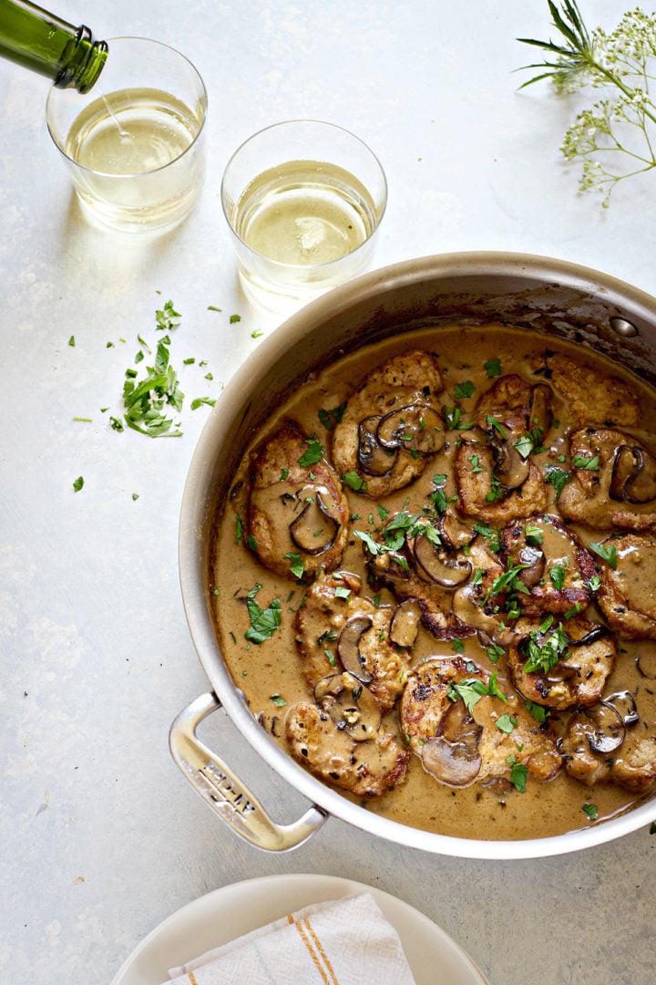 Top-down view of a skillet filled with pork marsala with mushrooms. Golden brown pork medallions are coated in a creamy brown marsala sauce with sliced mushrooms and sprinkled with chopped parsley. Two glasses of white wine sit in the background, one being poured. A scattering of fresh herbs is visible on the surface beside the skillet, and a folded dish towel rests on a plate at the bottom of the frame.