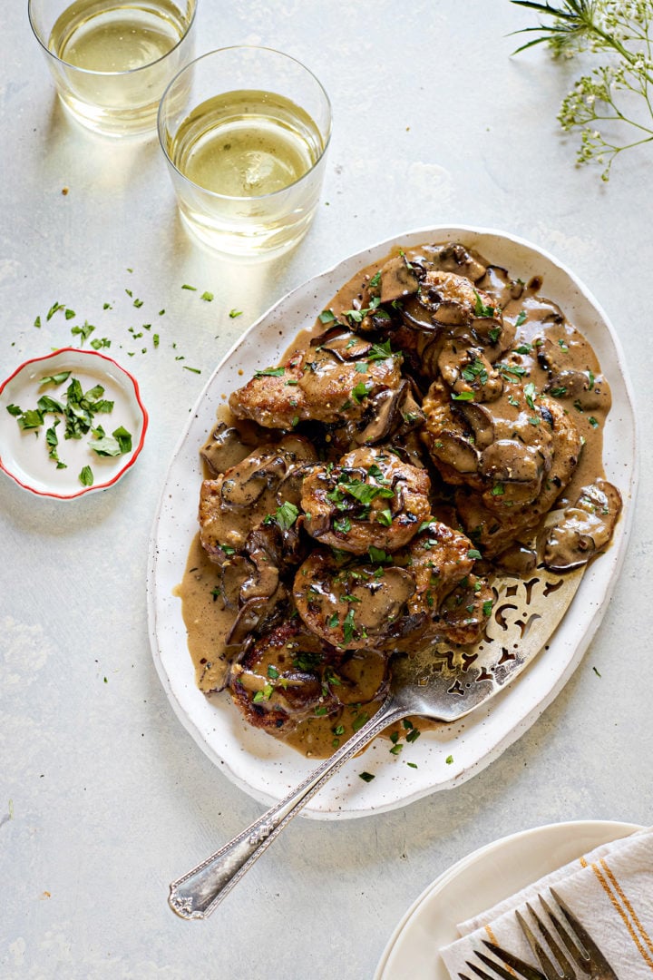 Overhead view of a serving platter filled with pork marsala recipe. The seared pork medallions are covered in a creamy mushroom sauce and sprinkled with chopped parsley. A decorative silver serving spoon rests on the platter. Two glasses of white wine, a small dish of extra parsley, and a plate with napkins and forks are visible around the edges of the scene.