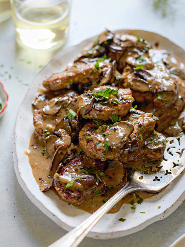 Overhead view of a serving platter filled with seared pork medallions topped with marsala sauce for pork. The creamy mushroom sauce is generously spooned over the meat and garnished with chopped parsley. A silver serving spoon rests on the platter. Two glasses of white wine and a small dish of additional parsley sit nearby on the light-colored surface.