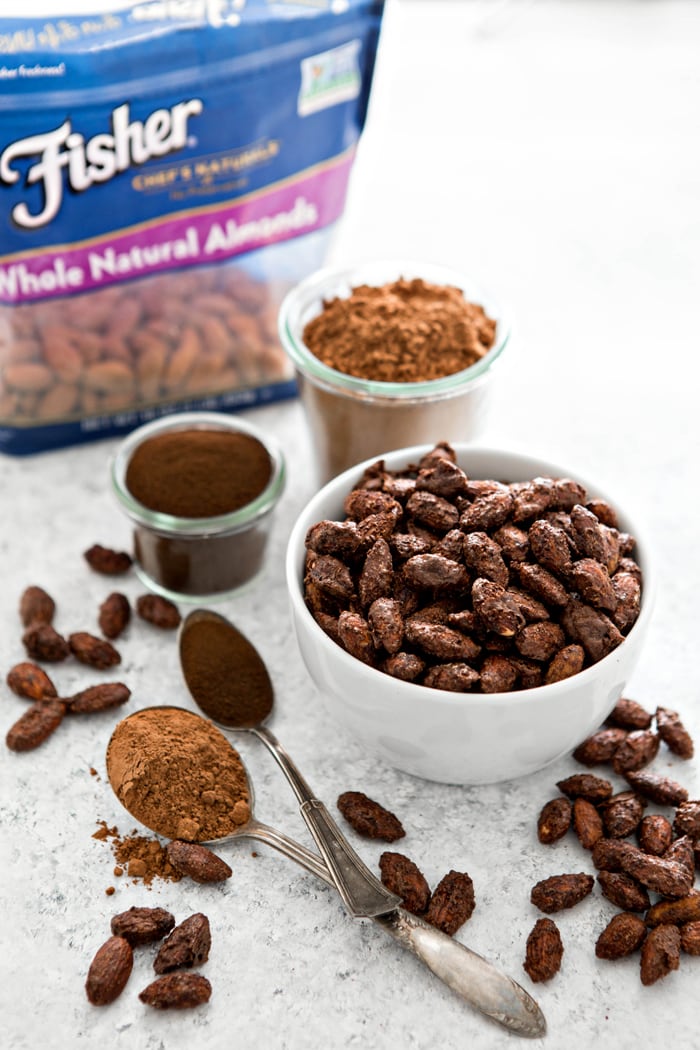 mocha roasted almonds in white bowl next to cocoa powder, espresso powder, and bag of nuts