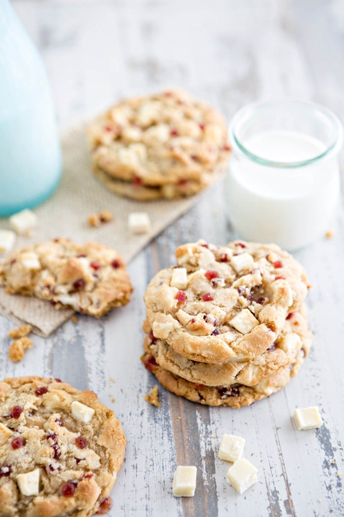 A stack of soft, chewy white chocolate raspberry cheesecake cookies with jammy raspberry bits and melty white chocolate chunks, served with a glass of milk on a rustic wooden surface.

