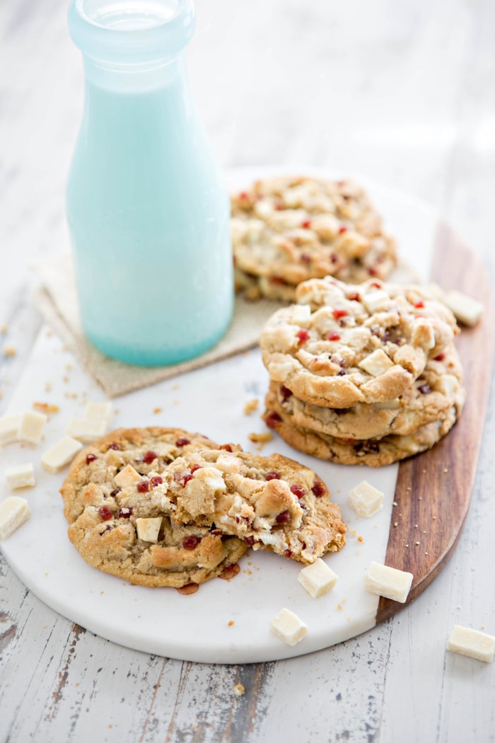 A batch of Subway-style white chocolate raspberry cheesecake cookies on a marble serving board, with a glass milk bottle and scattered white chocolate chunks.